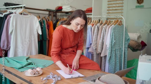 Woman fashion designer drawing sketch of new fashionable dress, sitting on table at workplace with laptop and sewing tools at atelier or workshop. Female dressmaker working in studio photo