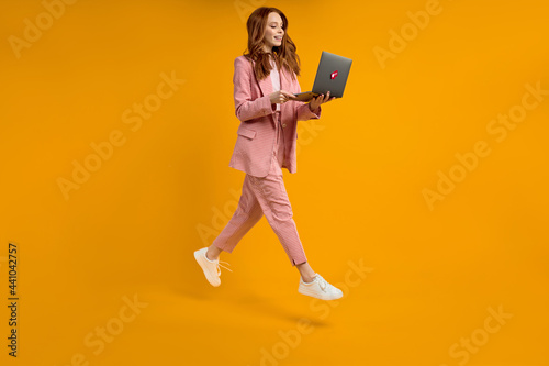 Red haired woman run jump typing laptop wearing elegant pink suit isolated on yellow background in studio, hurry up. side view portrait of lady working on laptop. Copy space for advertisement.