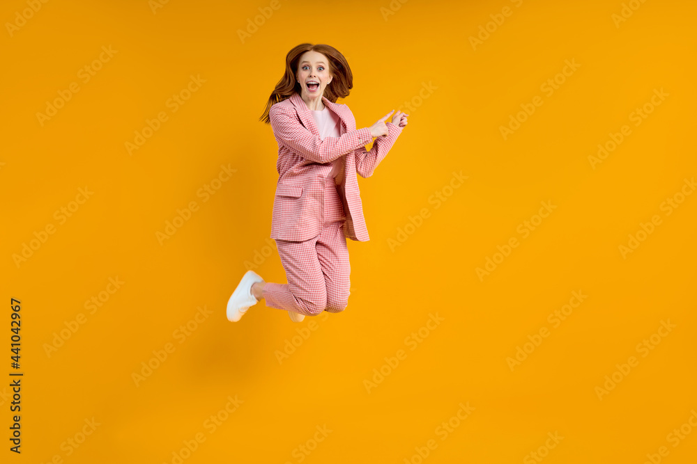 Full length photo of funny caucasian lady in pink suit jumping up high see low shopping prices direct finger at side on advert banner, isolated yellow color background in studio
