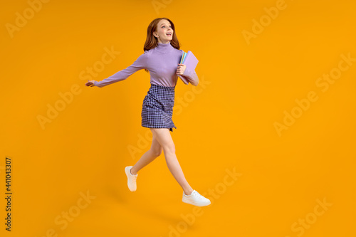 beautiful redhead lady at weekend jumping, hold notebooks in hands on way home, in air, wearing casual shirt and skirt isolated yellow orange background. Side view, copy space. Full-length portrait