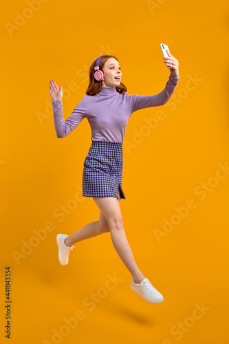 Excited Redhead Woman In Heaphones Jumping, Walking And Talking On Phone Online Via Camera, Say Hello, Smiling, Isolated On Yellow