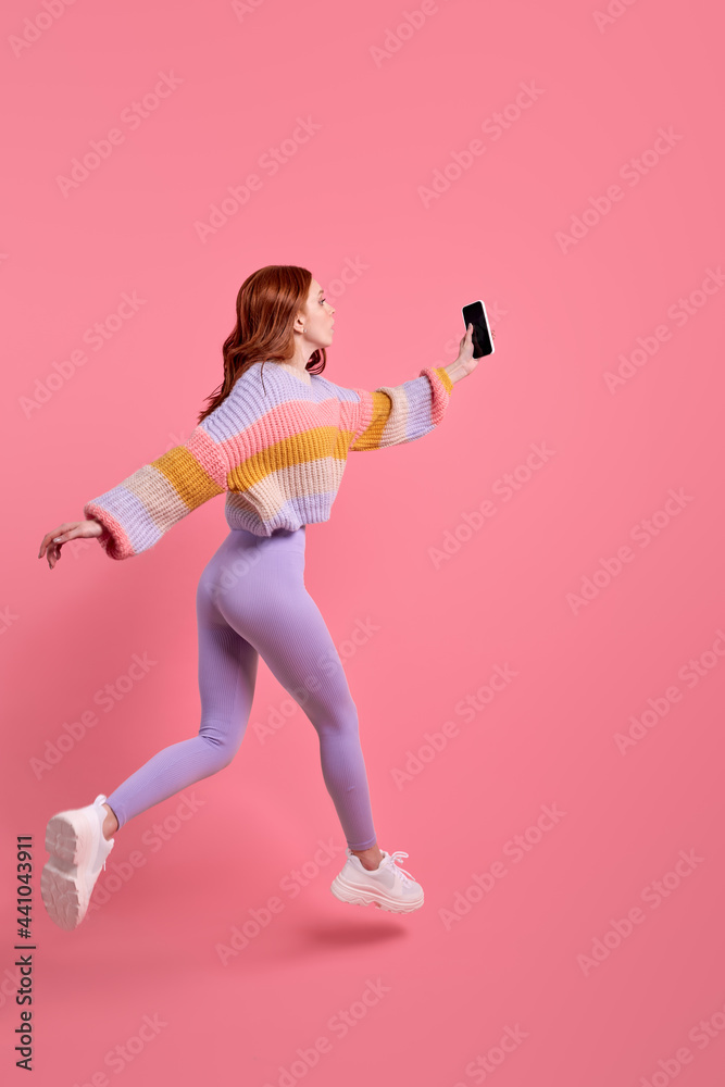 side view portrait of redhead female in leggins running jumping forward with smartphone in hands, taking photo of something, hurry. isolated pink background