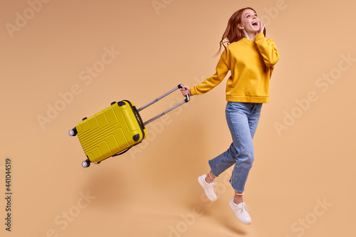 Traveler tourist redhead woman with suitcase, jumping in casual outfit isolated in studio. Caucasian Female passenger traveling abroad on weekends. Air flight journey concept. Copy space photo