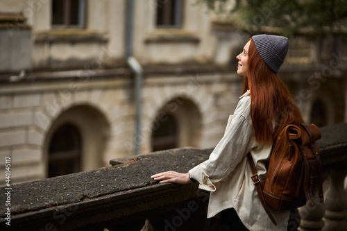 Slim graceful redhead woman in coat with backpack standing in old building on balcony, female is looking away with smile, enjoying attractions. Side view portrait