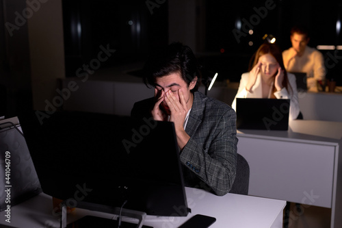 Foto young caucasian tired brunnete man in suit sits at night in office, eyes hurt from overexertion, suffering from headache