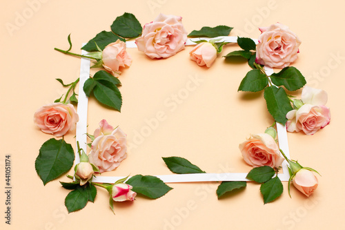 small white and pink flowers of a rose with green young leaves lie around a white empty frame on a pastel beige background. Floral minimal mockup template concept for spring holidays