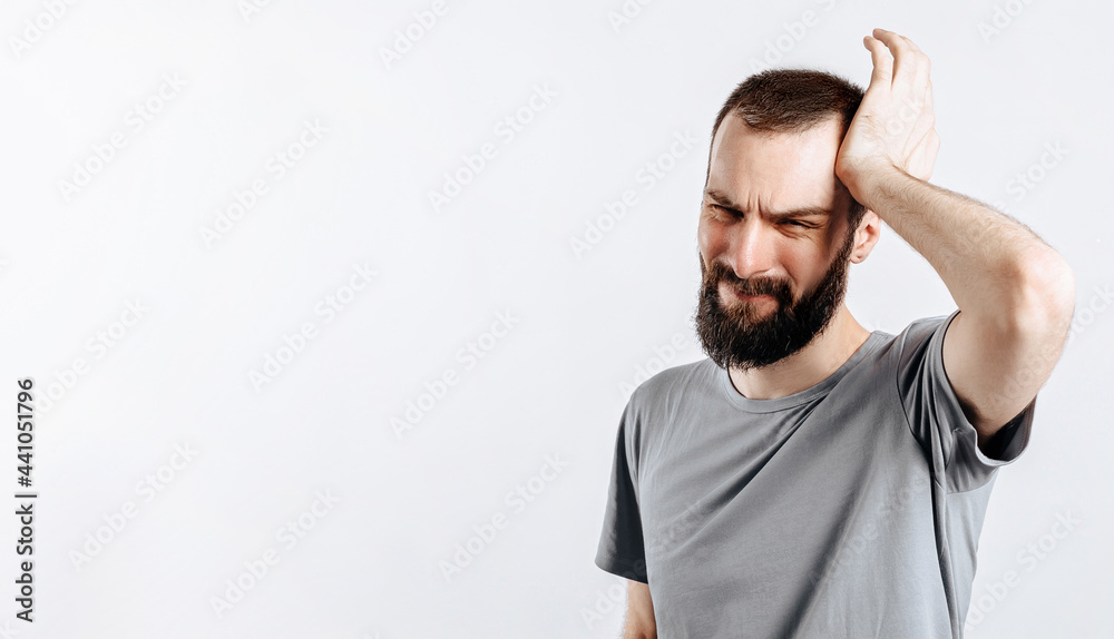 Naklejka premium Portrait of handsome young man frowning while holding hands near head on white background with space for advertising mock up