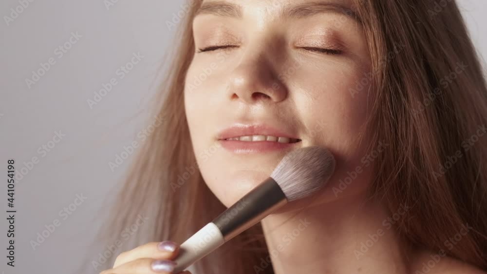 Natural makeup. Decorative cosmetics. Beauty care. Happy smiling brunette woman enjoying touching radiant face skin with soft powder brush isolated on beige copy space background. Natural makeup. Decorative cosmetics. Beauty care. Happy smiling brunette woman enjoying touching radiant face skin with soft powder brush isolated on beige copy space background.