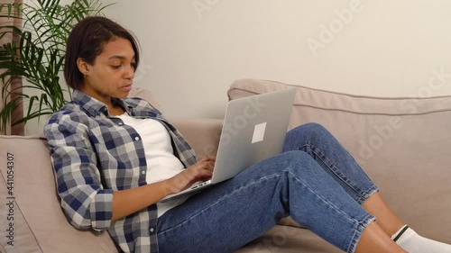 Beautiful young female freelancer working at home online lying down with a laptop. Checkered shirt, jeans. 4k video.