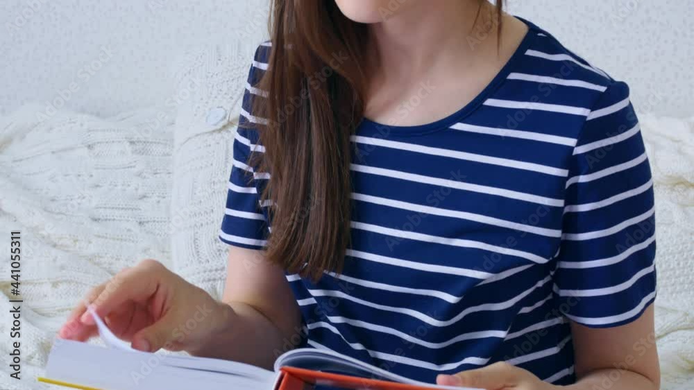 The unrecognizable dark-haired woman flips through the pages of the book, holding the open book at a considerable distance from her eyes. The woman is sitting on a knitted bedspread. Homeschooling. The unrecognizable dark-haired woman flips through the pages of the book, holding the open book at a considerable distance from her eyes. The woman is sitting on a knitted bedspread. Homeschooling.