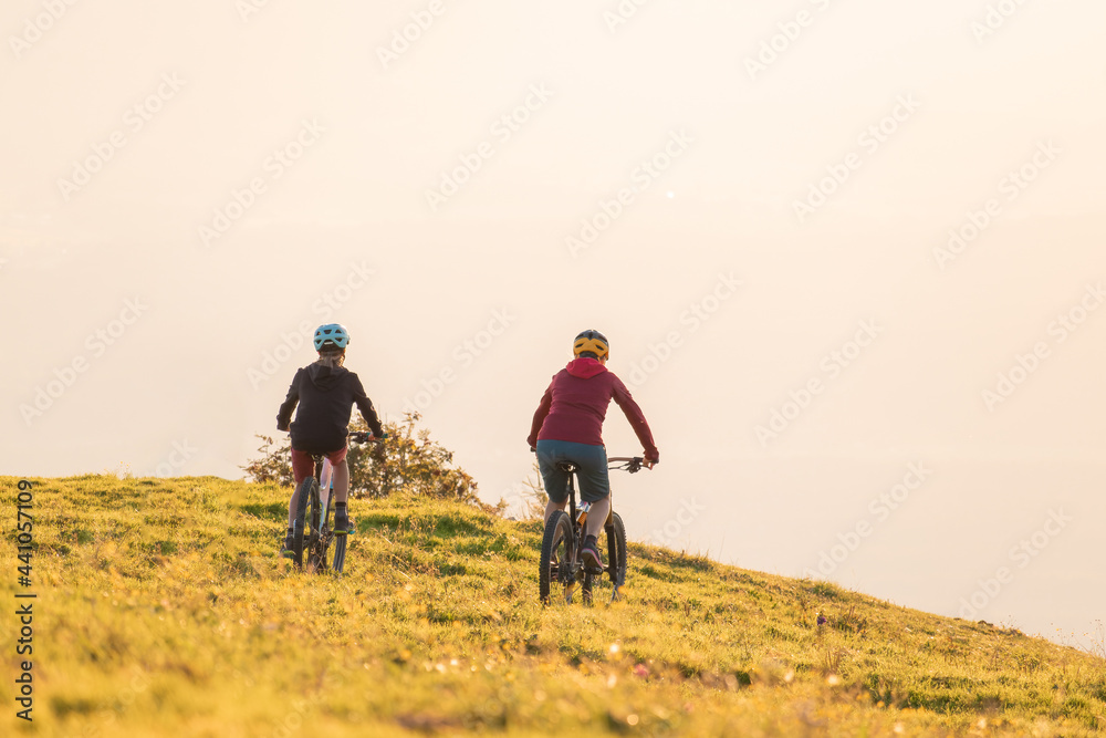 Fototapeta premium Mother and daughter cycling uphill with mountain bikes at a sunset.