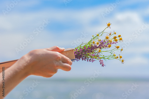 female hand holding a book with a bouquet of wildflowers against the background of the sea and sky