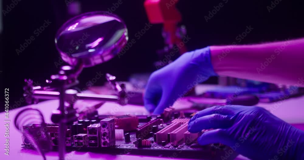 Close-up of assembling motherboard in neon light, fixing details for gadget in protective blue gloves. Repairing computer components with magnifying glass and screwdriver at electronic devices factory Close-up of assembling motherboard in neon light, fixing details for gadget in protective blue gloves. Repairing computer components with magnifying glass and screwdriver at electronic devices factory