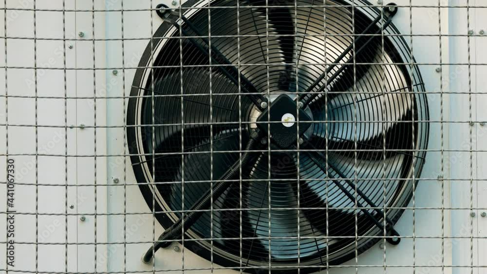 Industrial refrigeration unit mounted on wall of warehouse building. Unit with external cooling fans. Rotating blades. Metal turbine. Air conditioning equipment. Protective steel grill. Freezer unit