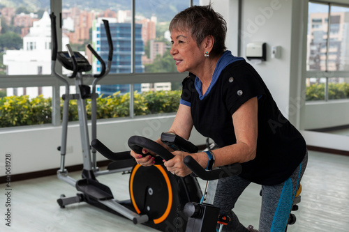 72-year-old grandmother works out in a gym doing weight routines and practicing exercise on a stationary bike.