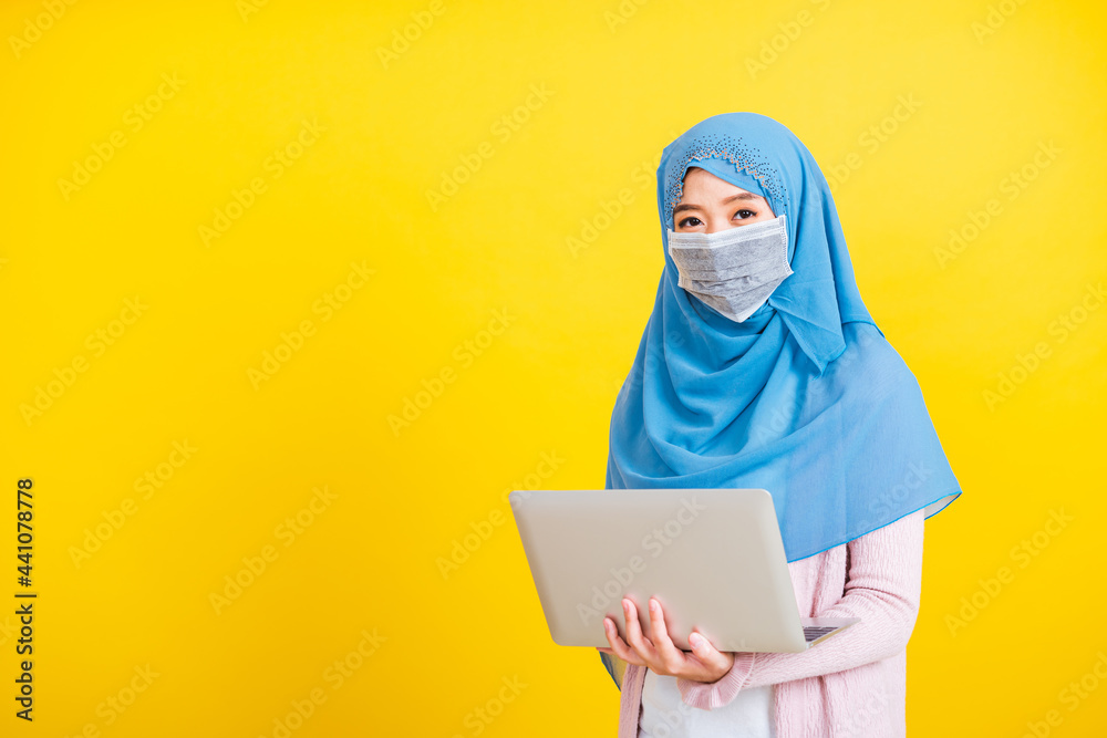 Asian Muslim Arab, Portrait of happy beautiful young woman religious wear veil hijab and face mask protective to prevent coronavirus she holding the laptop typing, isolated on yellow background Asian Muslim Arab, Portrait of happy beautiful young woman religious wear veil hijab and face mask protective to prevent coronavirus she holding the laptop typing, isolated on yellow background
