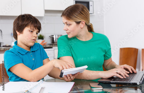 Annoyed mother helping son with homework sitting nearby at kitchen table