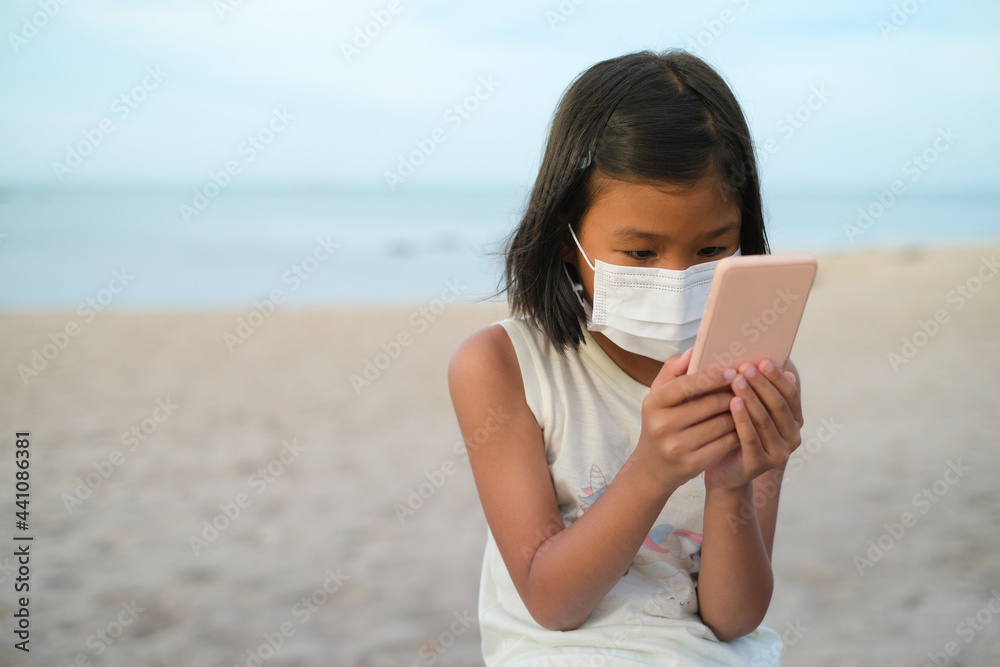 Asian girl with wearing protective face mask playing the smartphone at the beach with blurred background. Children new generation addicted with modern technology concept.