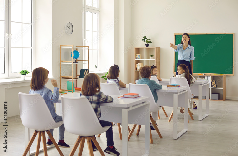 Little student raises hand and answers teacher's question in class. Happy female teacher asks kids questions during lesson in modern classroom interior. Concept of elementary school education Little student raises hand and answers teacher's question in class. Happy female teacher asks kids questions during lesson in modern classroom interior. Concept of elementary school education
