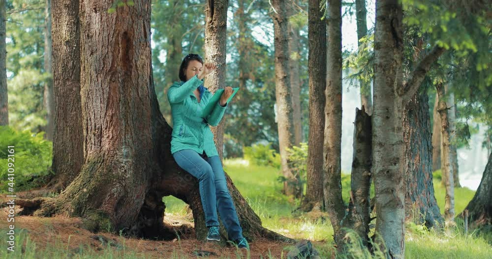 A girl draws in the forest. She is sitting on the root of a large tree and holds a paper stand in her hand. She has a pencil in her right hand. A girl draws a landscape. A girl draws in the forest. She is sitting on the root of a large tree and holds a paper stand in her hand. She has a pencil in her right hand. A girl draws a landscape.