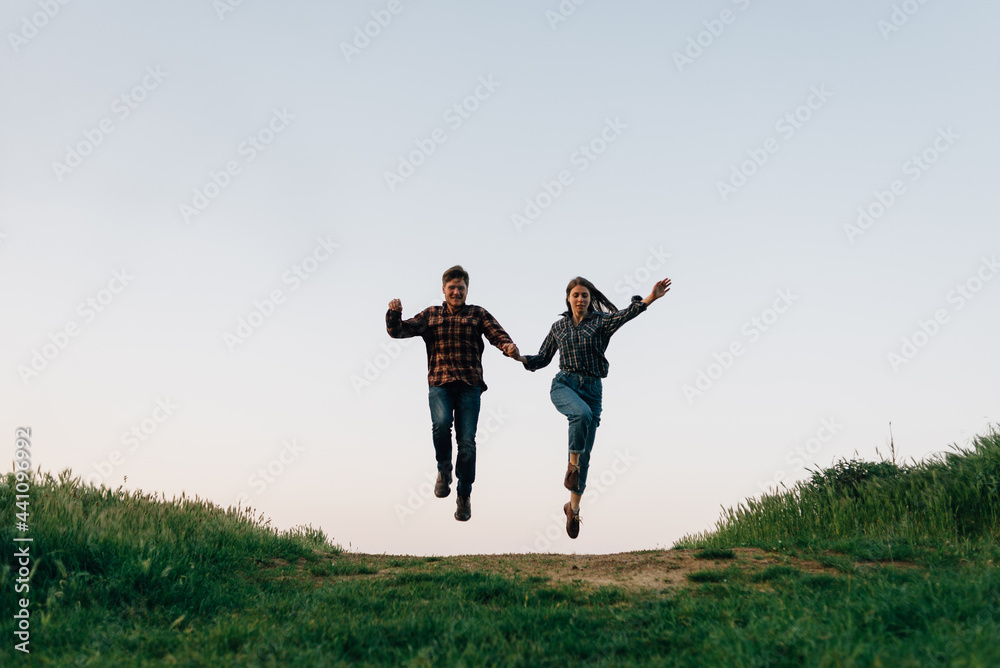 happy couple family man and woman jumping on the field road on the background blue sky. The guy and the girl are jumping holding hands while walking along a rural road. happy couple family man and woman jumping on the field road on the background blue sky. The guy and the girl are jumping holding hands while walking along a rural road.