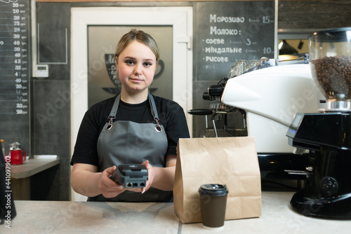 A barista woman in a cafe holds a payment terminal with nfs to pay for an order in a paper bag and a hot morning espresso coffee. Workplace coffee drinks seller.