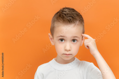 Little boy points a finger to his head isolated on an orange background. Concept of great idea or thought, good memory. 