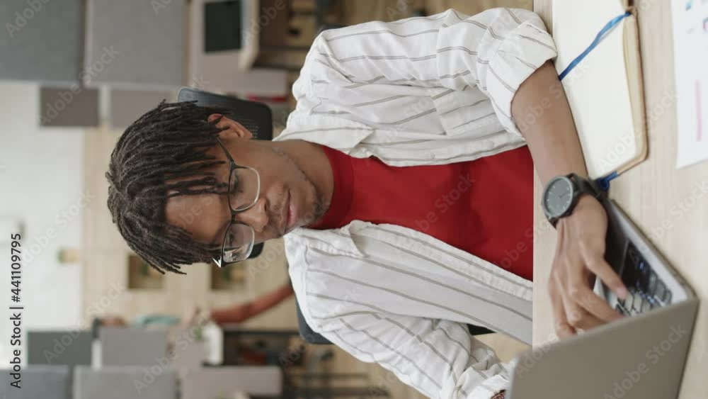 Vertical view shot of young African-American man with dreads wearing casual clothes sitting at desk in office, working on computer then looking at camera and smiling Vertical view shot of young African-American man with dreads wearing casual clothes sitting at desk in office, working on computer then looking at camera and smiling