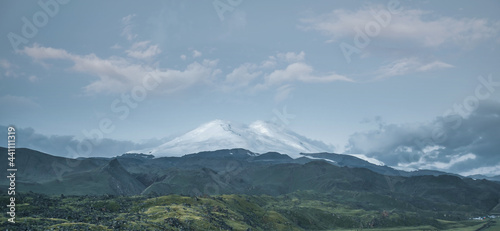 Natural landscape with snowy peaks of Mount Elbrus and a hilly green valley at sunrise.
