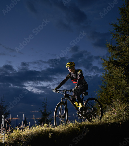 Smiling young man riding bicycle downhill with beautiful blue evening sky on background. Bicyclist in sports cycling suit cycling down grassy hill at night. Concept of sport, biking and active leisure