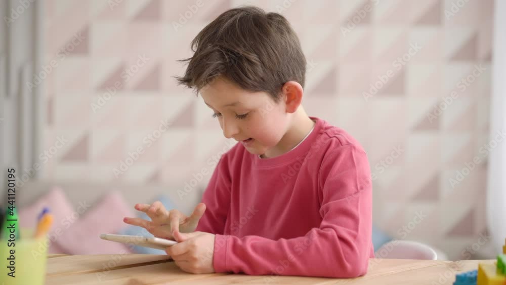 Funny schoolboy boy using tablet computer, sitting at home alone, positive child kid looking at gadjet screen, playing video game, watching cartoons, browsing apps, having fun with device Funny schoolboy boy using tablet computer, sitting at home alone, positive child kid looking at gadjet screen, playing video game, watching cartoons, browsing apps, having fun with device