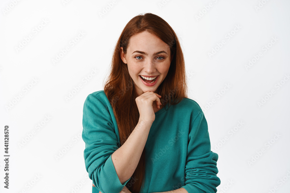 Portrait of cheerful young woman with red hair, listening something interesting, touch chin thoughtful, laughing and smiling, white background Portrait of cheerful young woman with red hair, listening something interesting, touch chin thoughtful, laughing and smiling, white background