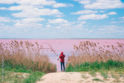 Man's figure in orange jacket with phone in hands, who is standing on the back of pink lake among dry reed and green grass. A man is capturing pink water with his phone.