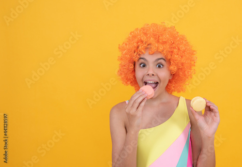 happy kid in swimsuit wearing orange curly wig hair eating french macaron, dental care.