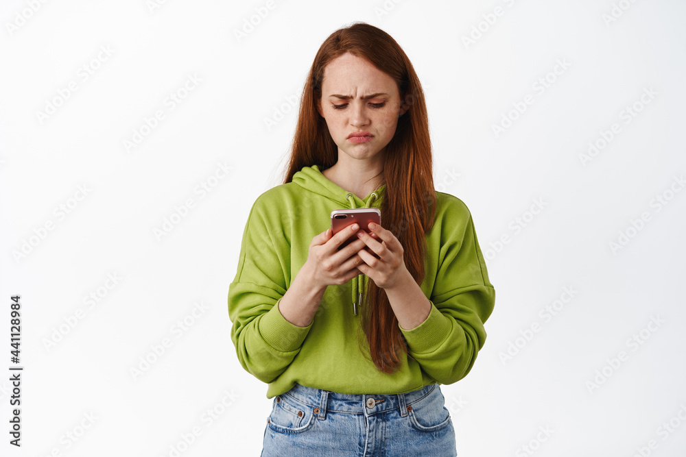 Sad and disappointed ginger teen girl looking upset at her phone, staring at smartphone with regret or jealousy, standing against white background Sad and disappointed ginger teen girl looking upset at her phone, staring at smartphone with regret or jealousy, standing against white background