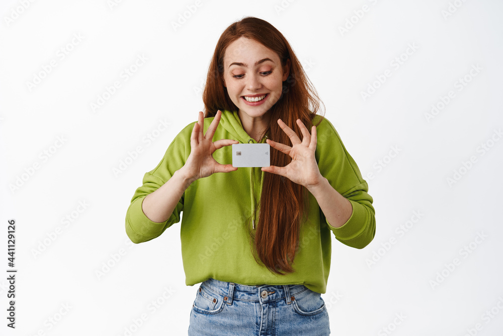 Smiling redhead girl looking at credit card, shopping contactless, making purchase, standing in casual outfit against white background Smiling redhead girl looking at credit card, shopping contactless, making purchase, standing in casual outfit against white background