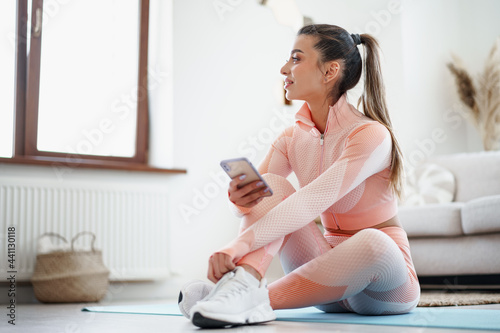 Beautiful young woman in sportswear sitting indoors and looking at smartphone before workout