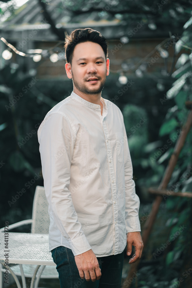 Fototapeta premium Portrait of a casually dressed handsome young Asian man smiling while standing alone outside on garden.