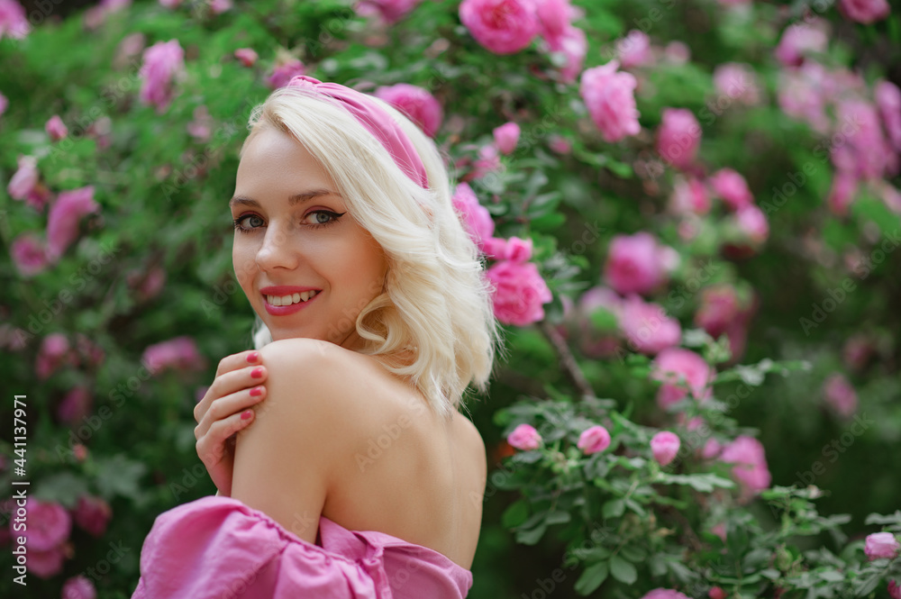 Happy smiling beautiful blonde woman wearing pink headband posing in blooming rose garden. Copy, empty space for text Happy smiling beautiful blonde woman wearing pink headband posing in blooming rose garden. Copy, empty space for text