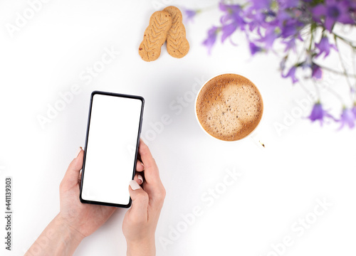 A cup of coffee, two cookies, bell flowers, and a phone in a woman's hands with a blank white screen. Top view, space for text.