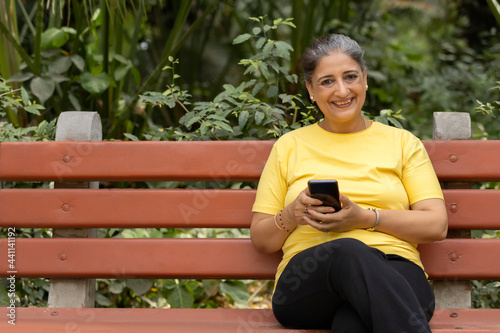 A CHEERFUL ADULT WOMAN SITTING ON A PARK BENCH HOLDING MOBILE PHONE