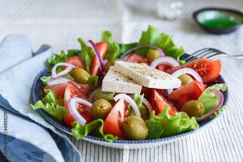Greek salad of fresh vegetable with tomatoes, lettuce, olives, red onion and feta cheese in bowl on white striped linen tablecloth. Selective focus