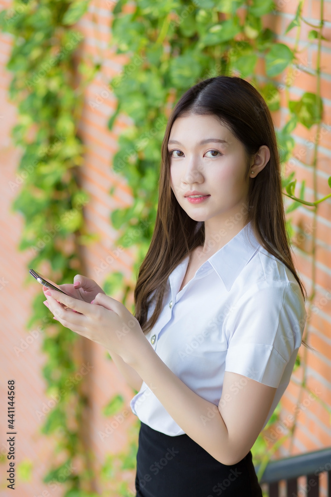 Portrait of an Asian Thai female student in a uniform is smiling happily while using a smartphone at university with trees and walls as a background. Portrait of an Asian Thai female student in a uniform is smiling happily while using a smartphone at university with trees and walls as a background.