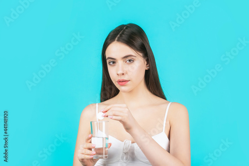 Girl take some pills, holds glass of water, isolated on blue. Young woman taking pill against headache. Brunette taking a pill with a glass of water. Woman taking drugs to releave headache photo