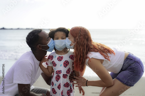 Multiethnic family kissing next to the sea while wearing a face mask that protects them from infection because of the outbreak of virus infection COVID-19. photo