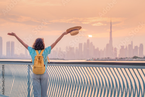 A happy traveler woman with a hat and a yellow backpack enjoys a stunning panoramic view of the Dubai Creek Canal and the famous tallest skyscraper Burj Khalifa
