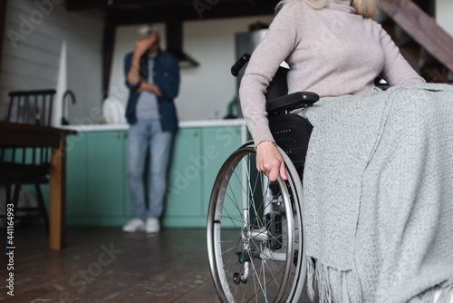 Senior woman sitting in wheelchair near husband in kitchen on blurred background