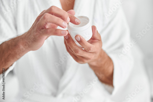 Man in bathrobe takes cream from open jar on light background closeup