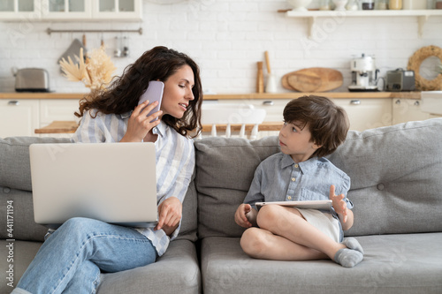 Multitasking casual businesswoman and mother work from home talk to kid, make phone call use laptop sit on couch in kitchen. Modern mom remote worker or freelancer lifestyle during covid-19 lockdown