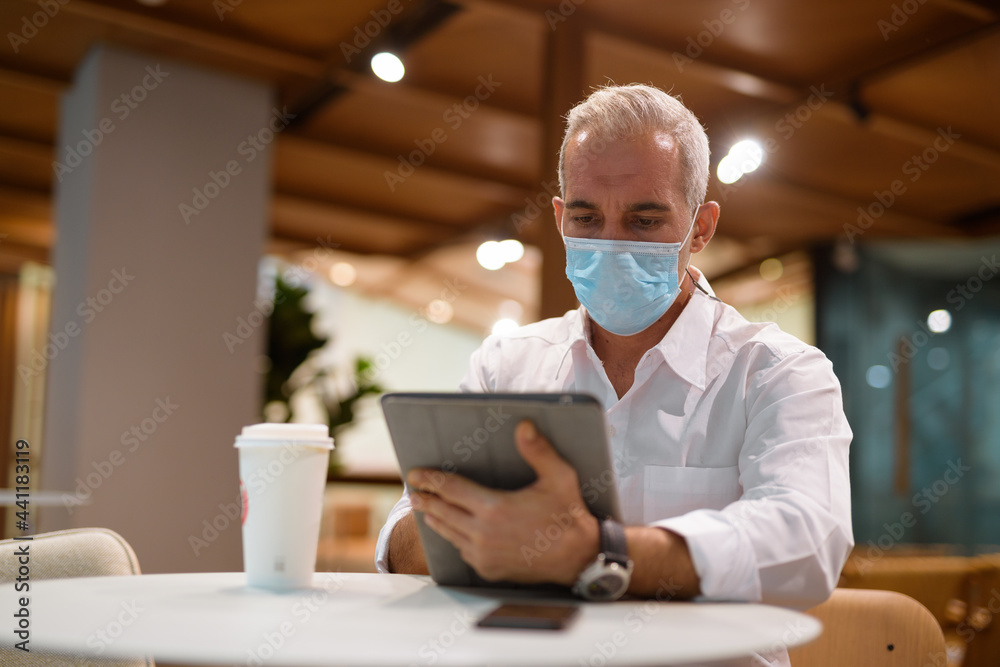 Businessman sitting at coffee shop and using digital tablet while wearing face mask and social distancing Businessman sitting at coffee shop and using digital tablet while wearing face mask and social distancing